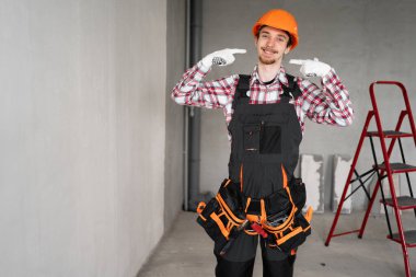 Erkek işçi, miğfer ve alet kemeri takan tamirci ya da tamirci inşaat alanında parmaklarıyla gülen dişe işaret ederek onarmaya ya da bina yapmaya hazırdır. Boşluğu kopyala