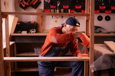 Meslek, sanat ve hobi. Yaşlı marangoz mavi tulum giyip garajda ahşap matkap kullanarak ahşap raf monte ediyor. Otantik atölye. Boşluğu kopyala