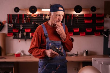 Düşünceli, olgun bir marangoz elinde elektrikli matkapla atölyede mobilya yenilemek için yeni adımlar planlıyor. El yapımı, küçük işletme, DIY ve girişimcilik konsepti.