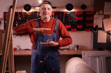 Olgun marangoz, atölyede mobilya yenileme projesinin boyutlarını gösteren büyük bir jest sergiliyor. El yapımı, küçük işletme, DIY ve ambalaj kavramı.