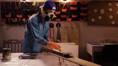 Marangoz, akşam garajda çalışan koruyucu maske ve eldiven takarak siyah renkli ahşap bir kalas boyuyor.