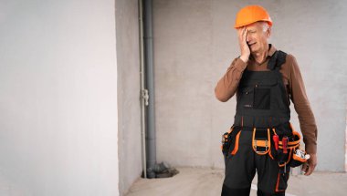 İnşaat işçisi ya da güvenlik miğferi takan tamirci, kızgın, stresli, tadilat sırasında inşaat alanında yaşadığı sorunlar yüzünden yüzünü eliyle kapatmış. Daire tamiri