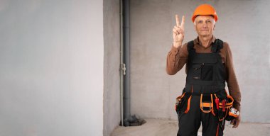 İnşaat işçisi ya da güvenlik miğferi takmış tamirci, tadilat sırasında inşaat alanında iki parmak zafer işareti gösteriyor. Daire tamiri ve yenileme konsepti. Pankart