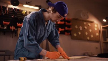 Küçük işletme konsepti. Genç marangoz mavi tulum giyip gece geç saatlerde garajda ya da ev atölyesinde ahşap tahta ölçüyordu. İş için alet kullanan profesyonel bir marangoz. Boşluğu kopyala