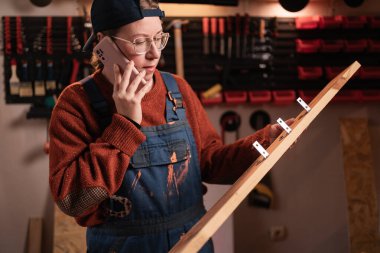 Bayan marangoz ya da zanaatkar müşteriyle akıllı telefondan konuşuyor, özel mobilya siparişini tartışıyor. Ahşap rafı tutan kadın küçük bir atölyede çalışıyor. Müşteri iletişimi kavramı.