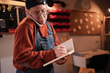 Kadın atölye çalışanı, akıllı telefondan müşteriyle konuşuyor, servis siparişini telefonla kabul ediyor, not defterine önemli notlar yazıyor. Müşteri desteği, el yapımı iş, servis kavramı.