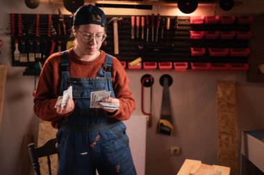 Tulum giyen kadın tamirci nakit parayı maaş olarak sayıyor, garajda ya da ev atölyesinde çalışırken banknot tutuyor, sıcak akşam ışığı, mavi yaka işi, gelir kavramı.