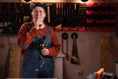 Tulumlu ve gözlüklü kadın tamirci tamirhanede çalışırken sessiz ve gizli bir işaret gösteriyor. Boşluğu kopyala