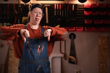 Tulumlu ve gözlüklü üzgün kadın tamirci tamirhanede çalışırken baş parmağının hoşlanmadığı hareketi gösteriyor. Boşluğu kopyala