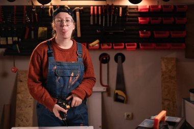 komik aptal kadın tamirci tulumlu ve gözlüklü tamirci tamirhanede çalışırken dilini gösteriyor. Boşluğu kopyala