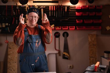 İş yerinde tamirat atölyesinde çalışırken umut dolu emojiler için dua eden işçi tulumlu ve gözlüklü kadın tamirci. Boşluğu kopyala