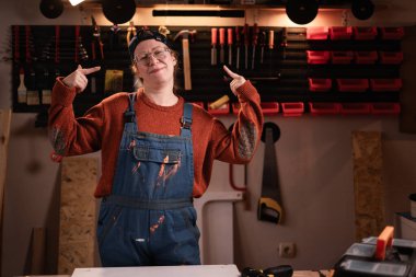 Tulumlu ve gözlüklü mutlu kadın tamirci atölyede çalışırken gülümseyen parmaklarıyla atölyeyi işaret ediyor. Boşluğu kopyala