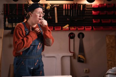 Tulumlu hasta kadın tamirci ve boğaz ağrısı ve grip için öksürük gösteren gözlüklü hasta kadın atölyede çalışırken atölyede soğuk algınlığı ya da alerji. Boşluğu kopyala