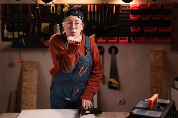 Kadın tamirci ve gözlüklü dudak büküyor ve tamirhanede çalışırken kameraya hava öpücüğü üflüyor. Boşluğu kopyala