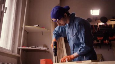 Profesyonel işçi tulumuyla elektrikli matkap kullanarak ahşap tahtada delik açıyor. Boşluğu kopyala