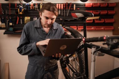 Dizüstü bilgisayarı olan genç bir tamirci. Tamir atölyesindeki iş adamı. Erkek bisiklet tamircisi dizüstü bilgisayarla çalışıyor. Boşluğu kopyala