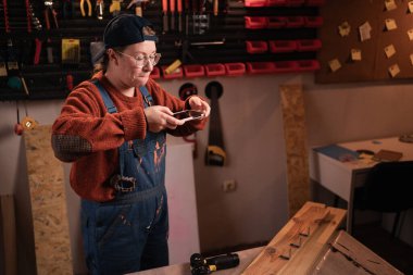 Garaj atölyesinde bitirilmiş ahşap projesinin fotoğrafını çekmek için akıllı telefon kullanan kadın marangoz. El yapımı mobilyaları belgeleyen profesyonel bir zanaatkar. Küçük işletme kavramı.