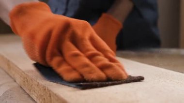 Turuncu eldivenli marangoz elleri zımpara kağıdıyla ahşap tahtayı zımparalıyor. yakın plan
