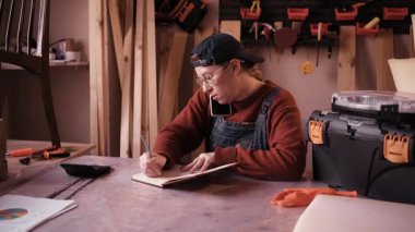 Profesyonel kadın marangoz müşteriyle telefonda konuşuyor ve atölyedeki not defterine sipariş detaylarını yazıyor. Küçük işletme yönetimi, müşteri hizmetleri ve zanaat girişimciliği kavramı.