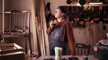 Kadın marangoz, modern marangozluk atölyesinde çalışma masasının yanında kahve molası veriyor. Çalışma alanı rahatlama, zanaatkar yaşam tarzı ve küçük işletme kavramı