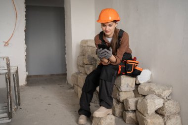 Miğferli kadın müteahhit ve yansıtıcı yelekli taş yığınının üzerinde oturuyor ve molada cep telefonu kullanıyor. Ev yenileme sahasında dijital haber okuyan profesyonel kadın müteahhit..
