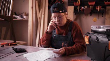 Atölyedeki stresli kadın marangoz, küçük marangoz işi için iflas risklerini yönetirken finansal sorunlar ve banka kredi başvurusu hakkında akıllı telefondan konuşuyor..