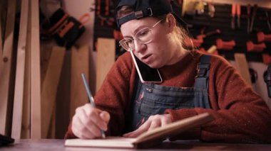 Profesyonel kadın marangoz atölyede oturmuş akıllı telefondan konuşuyor ve iş sırasında küçük marangozluk işlerini yönetirken müşteri siparişlerini not defterine yazıyor..