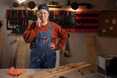 Profesyonel kadın marangoz garajında marangozluk aletleri kullanarak ahşapla çalışır. Ayakta duruyor ve telefonda konuşuyor. Otantik el sanatları atölyesi. El işleri, yaratıcı küçük işletmeler..
