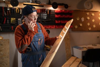 Profesyonel bir marangoz garajında marangozluk aletleri kullanarak ahşapla çalışır. Elinde bir tahtayla telefonda konuşuyordu. Otantik bir zanaat atölyesi.