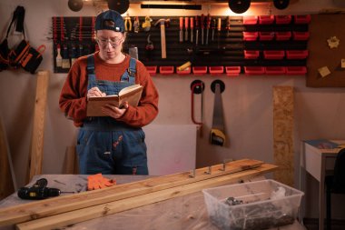 Profesyonel bir marangoz garajında marangozluk aletleri kullanarak ahşapla çalışır. Bir defter tutuyor ve notlar alıyor. El işi, yaratıcı küçük işletme.