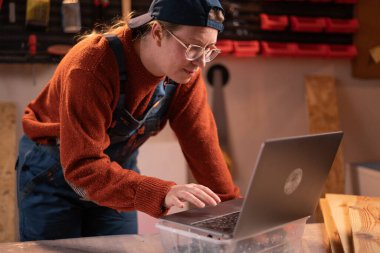 Profesyonel bir kadın marangoz garajında marangozluk aletleri kullanarak ahşapla çalışır. O bir masada duruyor ve bir dizüstü bilgisayar klavyesinde yazıyor. Mesleğin, sanatın ve hobinin kavramsallaştırılması.