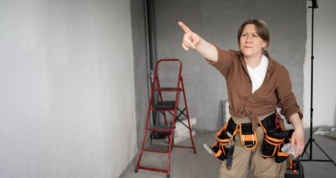 Ev dekorasyonunda çalışan, kameraya bakan ve mesaj için parmağıyla yeri gösteren alet kemeri takan bir kadın. Boşluğu kopyala