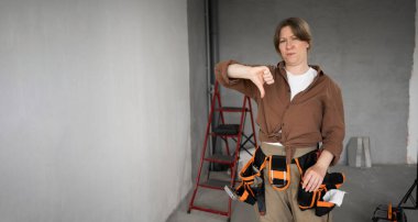 Mutsuz kadın tamirci ya da müteahhit alet kemeri takıyor, ev tadilatı sırasında baş parmak işareti gösteriyor. Kalitesiz iş kavramı, kötü inşaat hizmeti