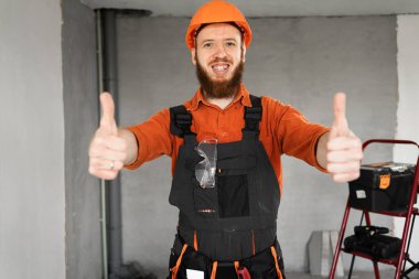 Meslek, inşaat ve inşaat kavramı. Sakallı erkek işçi ya da inşaatçı inşaat alanında ya da yeni dairede baş parmak gösteriyor. yakın plan