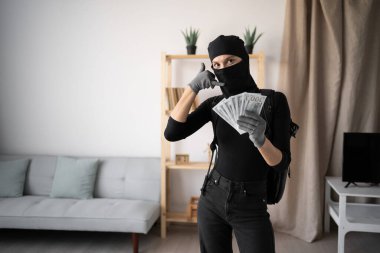 Siyah kar maskeli bir hırsız ya da soyguncu terörizm suçlusu evde para çalıyor banknotları gösteriyor ve kameraya bakarak telefon hareketi yapıyor. Boşluğu kopyala