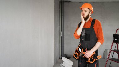 Sert şapkalı ve mavi tulumlu erkek sakallı müteahhit kemerinde diş ağrısı çeken ağız tutarak çalışıyor. Onarım, inşaat ve inşaat kavramı