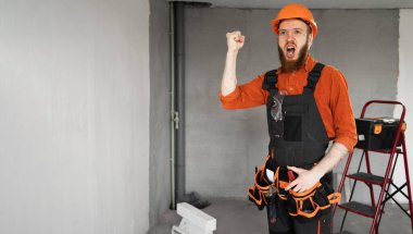 Sert şapkalı ve mavi tulumlu erkek sakallı müteahhit kemerinde iş aletleriyle agresif duygularla bağırıyor. Onarım, inşaat ve inşaat kavramı