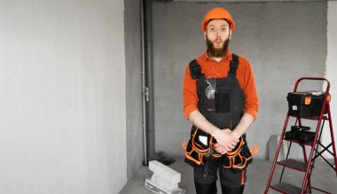 Sert şapkalı, mavi tulumlu, kemerinde iş aletleri olan erkek sakallı müteahhit balık dudaklı komik surat yapıyor. Onarım, inşaat ve inşaat kavramı