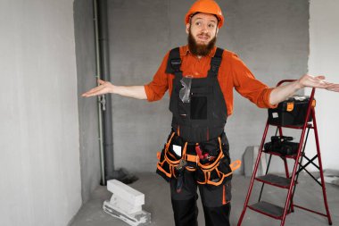 Sert şapkalı, mavi tulumlu, kemerinde iş aletleri olan erkek sakallı müteahhit şüphe götürmez bir jest yapıyor. Onarım, inşaat ve inşaat kavramı