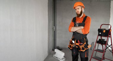 Sert şapkalı ve mavi tulumlu erkek sakallı müteahhit kemeriyle mutsuz bir şekilde kollarını kavuşturdu. Onarım, inşaat ve inşaat kavramı