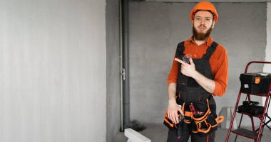 Sert şapkalı ve mavi tulumlu erkek sakallı müteahhit kemer ucu işaret parmağıyla bölgede çalışıyor. Onarım, inşaat ve inşaat konsepti