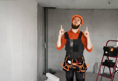 Kemerinde iş aletleri olan sert şapkalı ve mavi tulumlu erkek sakallı müteahhit fotokopi mekanını işaret ediyor. Onarım, inşaat ve inşaat konsepti