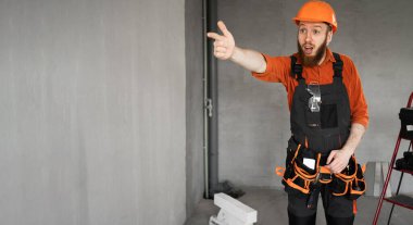 Sert şapkalı ve mavi tulumlu erkek sakallı müteahhit kemeriyle sana bakıyor ve kameraya bakıyor. Onarım, inşaat ve inşaat konsepti