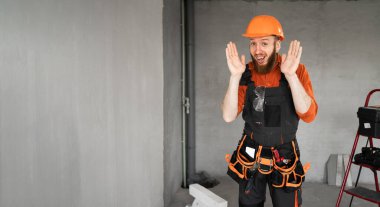 Sert şapkalı, mavi tulumlu, kemerinde iş aletleri olan ve elleri havada gülümseyen sakallı mutlu bir erkek inşaatçı. Onarım, inşaat ve inşaat konsepti