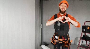 Kasketli ve mavi tulumlu mutlu erkek sakallı müteahhit kemerinde çalışan aletlerle kamerada sevgi dolu kalp hareketi gösteriyor. Onarım, inşaat ve inşaat konsepti