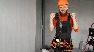 Kasketli ve mavi tulumlu mutlu erkek sakallı müteahhit kemeriyle zaferi kutluyor ya da yumruklarıyla zaferi kazanıyor. Onarım, inşaat ve inşaat konsepti