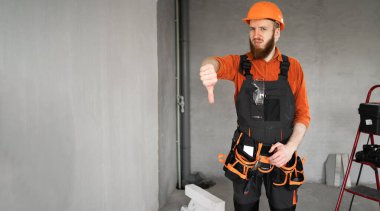 Mutsuz, sakallı, sert şapkalı, mavi tulumlu, kemerinde iş aletleri olan, hoşlanmadığı jestler gösteren erkek müteahhit. Onarım, inşaat ve inşaat konsepti