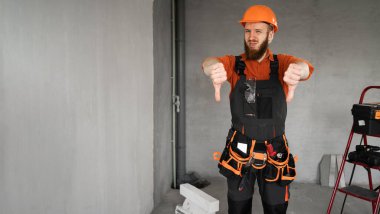 Mutsuz, sakallı, sert şapkalı, mavi tulumlu, kemerinde iş aletleri olan, hoşlanmadığı jestler gösteren erkek müteahhit. Onarım, inşaat ve inşaat konsepti