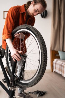 Turuncu gömlek giyen genç, uzun saçlı adam evde bisiklet tamir ediyor. Boşluğu kopyala