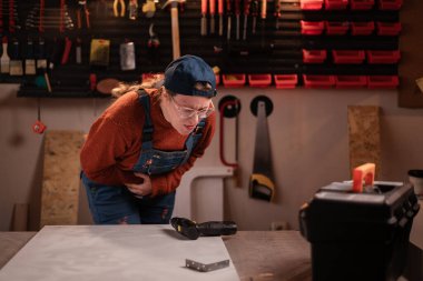 Sağlık sorunu kavramı. Kadın katılımcı atölye sahnesinde atölye kıyafeti giyer. Duvarda mide ağrısı çeken aletler asılı durur. Bayan marangoz gece geç saatlerde otantik garajda. Boşluğu kopyala